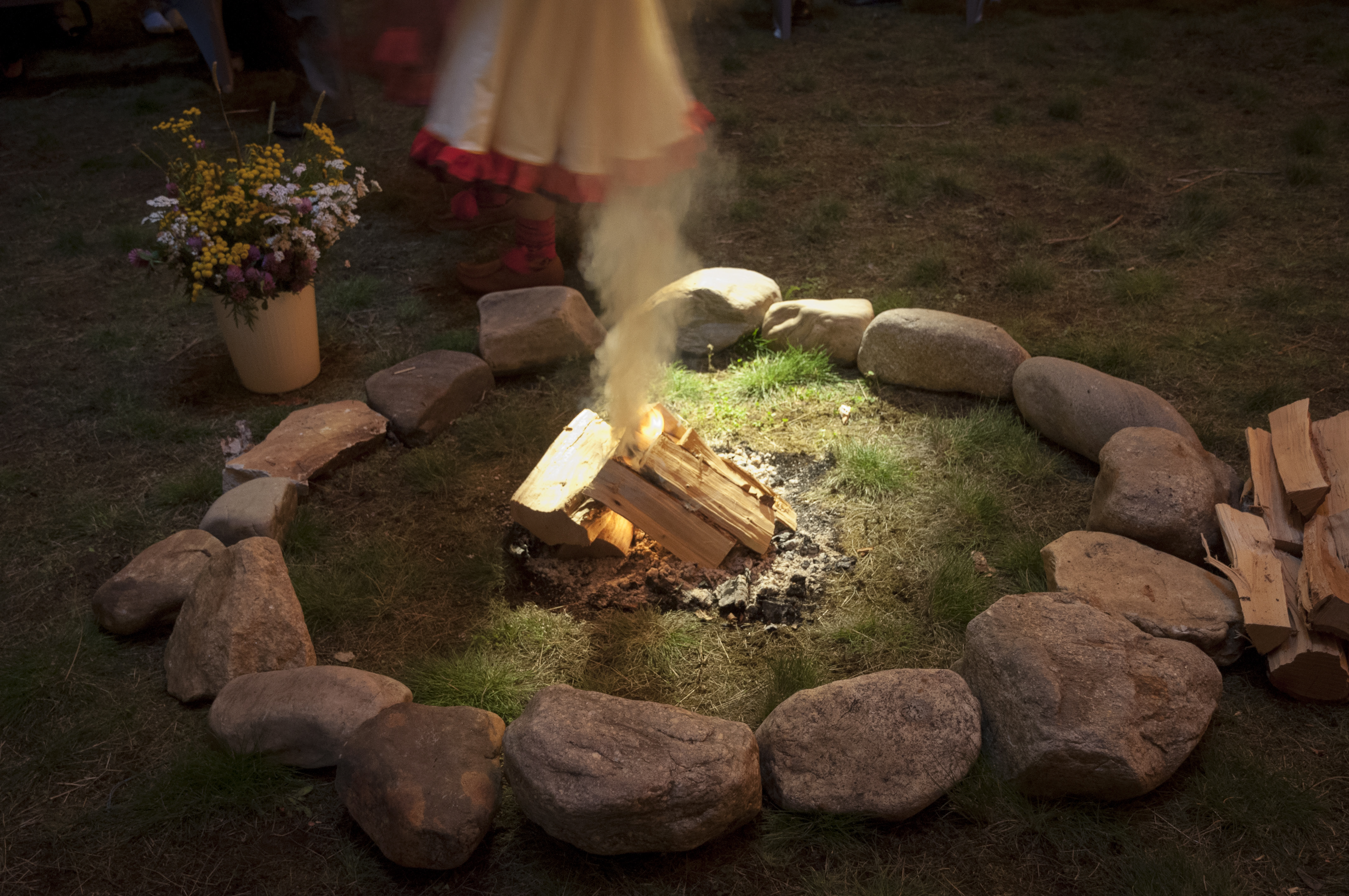 Någon med en samisk klänning och samiska skor står vid en eldstad med liten brasa och en hink med sommarblommor.