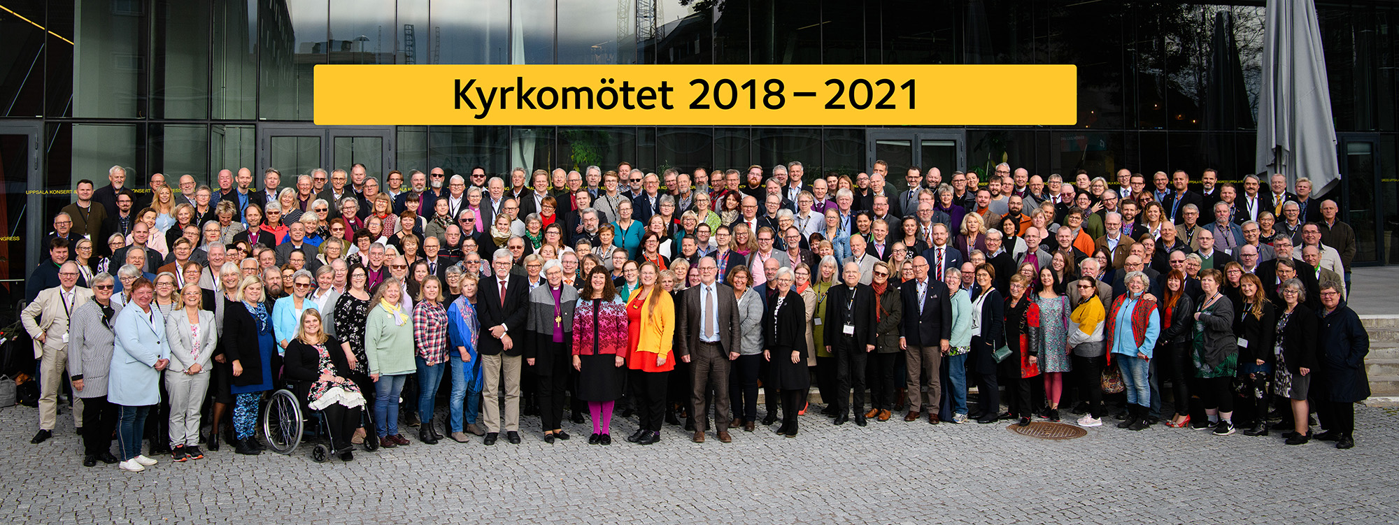 Kyrkomötets deltagare framför Uppsala konsert och kongress, med en skylt i bakgrund med texten: Kyrkomötet 2018-2022.