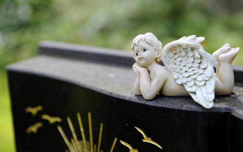 En ängel i sten ligger på mage på en gravsten, på gravstenen syns inristade fåglar och streck i guld.