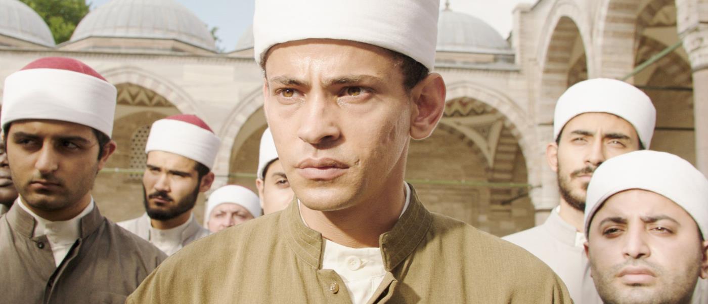 Foto från filmen där huvudpersonen står främst med ett antal män bakom sig, och en moskéliknande byggnad i bakgrunden.