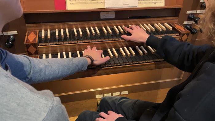 En organist och en orgelelev sitter ner tillsammans vid Helga Trefaldighets kyrkas orgel och spelar tillsammans på manualernas tangenter.