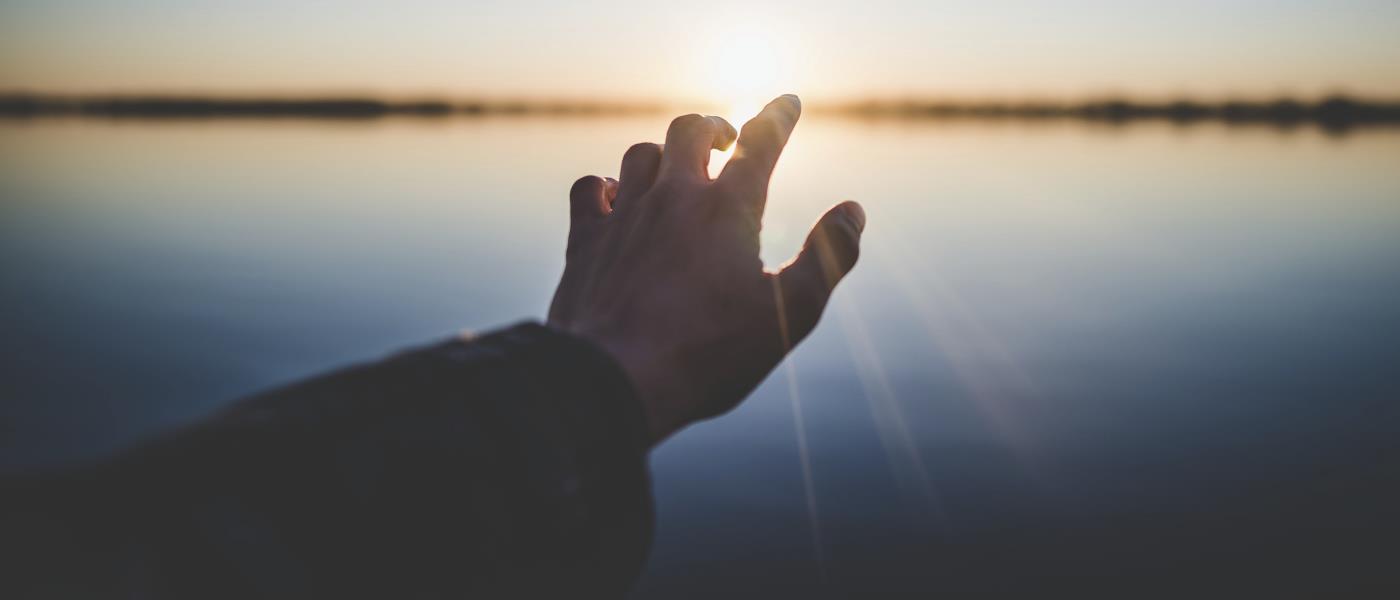 En människa sträcker ut sin hand mot solnedgången över ett vattendrag.