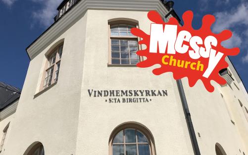 Vindhemskyrkans fasad syns mot en blå himmel och i bildens ena hörn syns också en färgglad skylt som har texten "Messy Church".