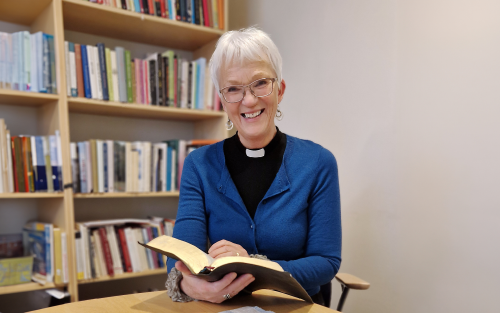 Leende kvinna med kort vitt hår, blå kofta och prästkrage runt halsen i svart skjorta sitter framför suddig bokhylla och håller i en bok