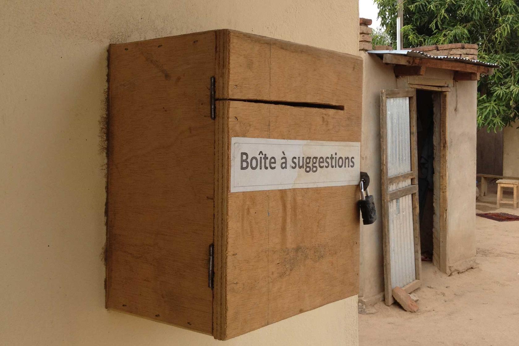 En låda med texten Boite a suggetions - förslagslåda sitter på en väg, i bakgrunden syns ett skjul.