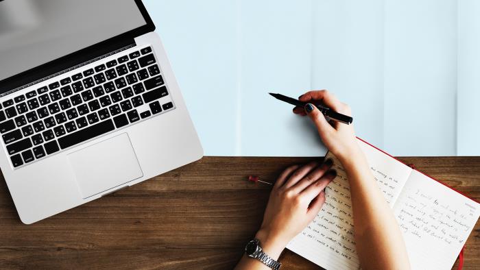 Bild ovanifrån på en dator, en kaffekopp, en hand som håller i en penna och en skrivbok.