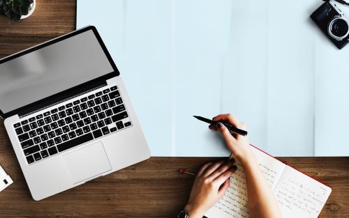 Bild ovanifrån på en dator, en kaffekopp, en hand som håller i en penna och en skrivbok.