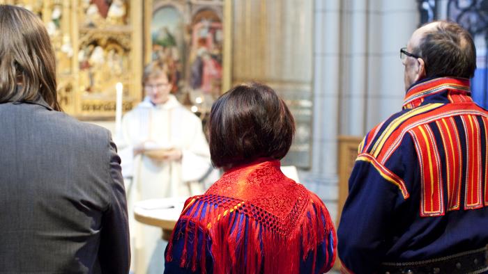 Tre personer varav två klädda i samiska folkdräkter står längst fram vid en gudstjänst i kyrkan.