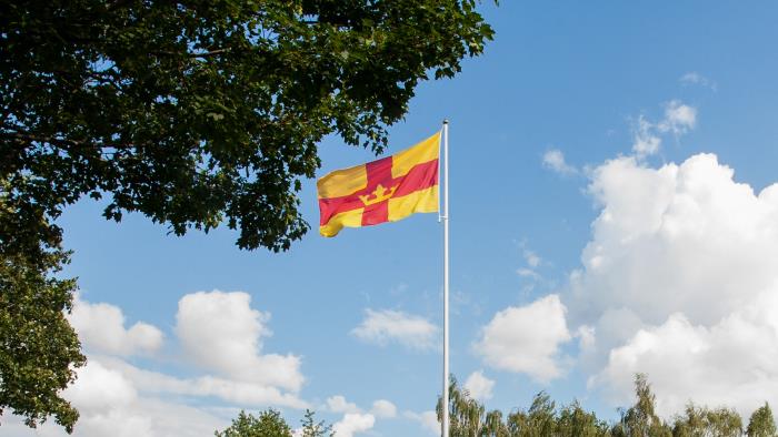 En flaggstång med Svenska kyrkans flagga mot blå himmel. Träd i bakgrunden.