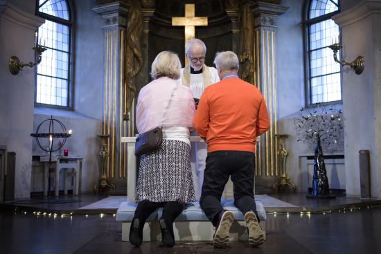 En man och en kvinna i vardagliga kläder knäböjer bredvid varandra framför en manlig präst i kyrkan.