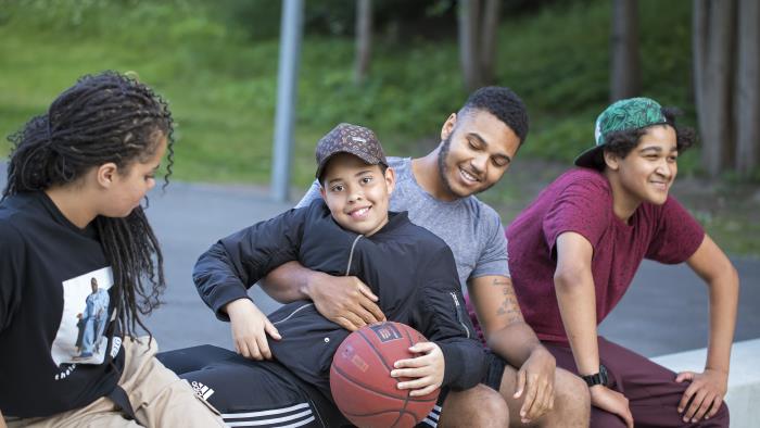 Några ungdomar sitter utomhus. En av dem kramar om en liten kille som håller i en basketboll.