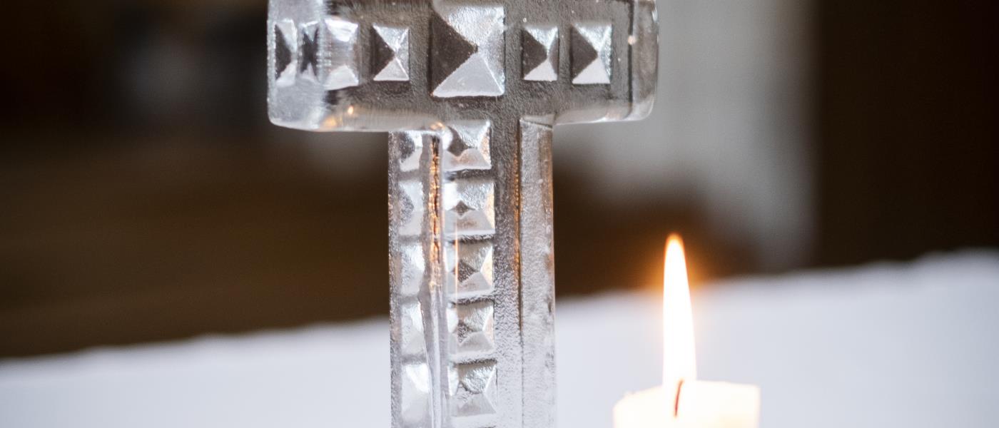 Ett kors av handgjort glas står på ett altare. Framför korset brinner ett ljus på en trefotad ljusstake.