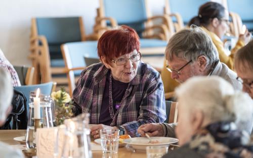 Några äldre personer sitter vid ett bord och äter soppa och samtalar.
