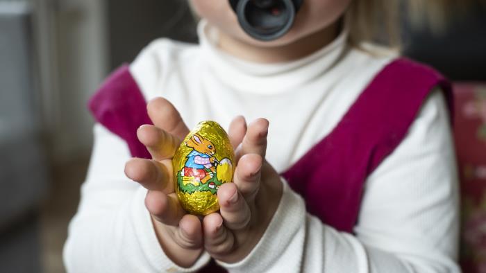 En liten flicka med napp håller upp ett påskägg i choklad. Det har foliepappret kvar.