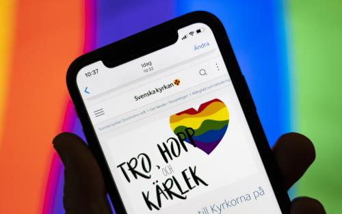Någon håller en mobiltelefon där skärmen visar Svenska kyrkans information om Pride. I bakgrunden syns regnbågsfärgerna.