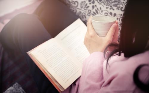 En kvinna sitter och läser med en bok i knät och en kopp i händerna.