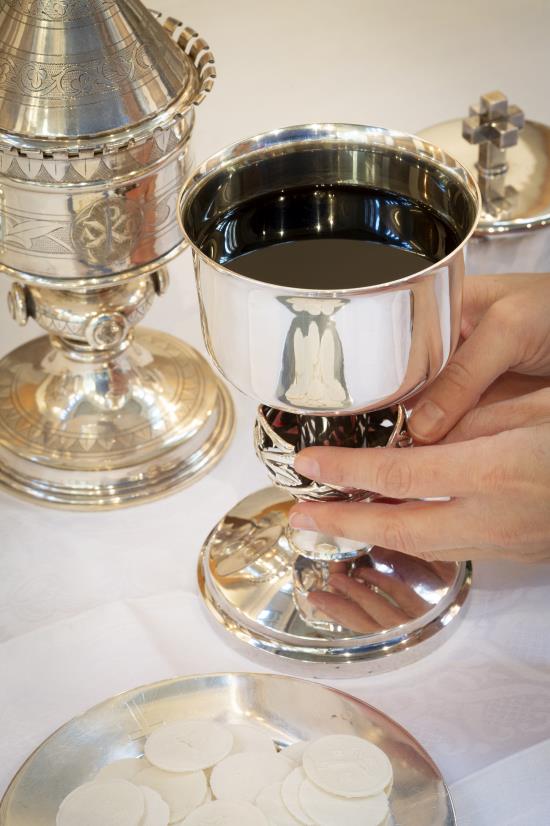 En person håller i en silverkalk fylld med nattvardsvin. Bredvid syns ett fat med oblater.