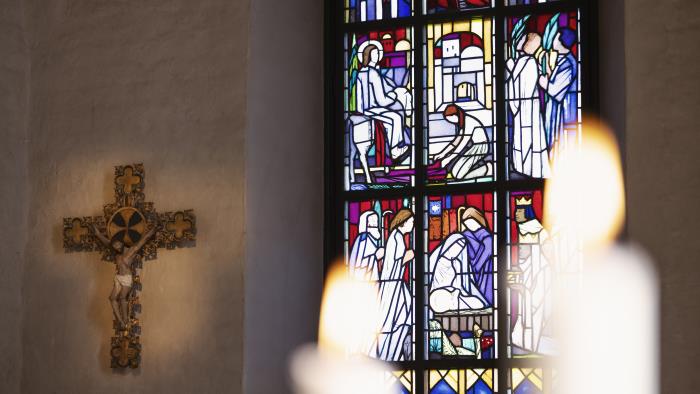 Ett krucifix hänger på väggen bredvid ett fönster med glasmålningar.