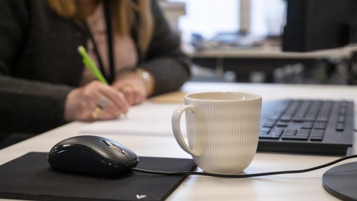 En kaffekopp står bredvid en datormus på ett skrivbord. En kvinna som antecknar syns suddigt i bakgrunden.