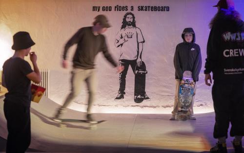 Ungdomar åker skateboard på en ramp. På väggen bakom är en målning och texten My god rides a skateboard.