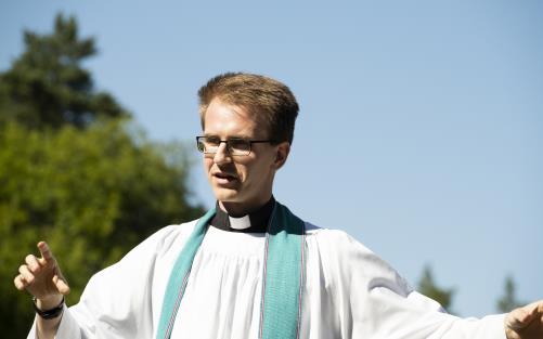 En manlig präst med vit kåpa och ljusblå stola står utomhus och gestikulerar med armarna.