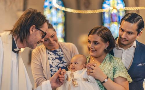 Ett dop i en kyrka. En kvinna med sin man bakom sig, håller barnet i famnen. En manlig präst håller barnets hand.