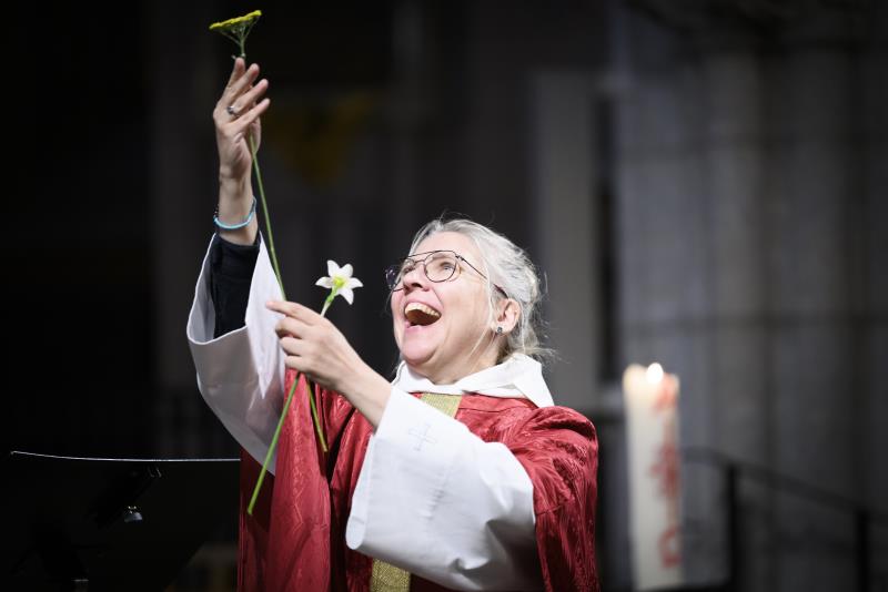 En kvinnlig präst står i en kyrka och håller leende upp två blommor mot taket.