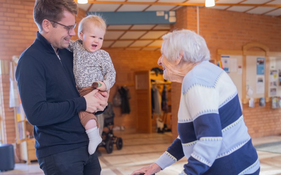 En äldre kvinna med rullator samtalar inomhus med en man och ett litet barn som han håller i famnen.