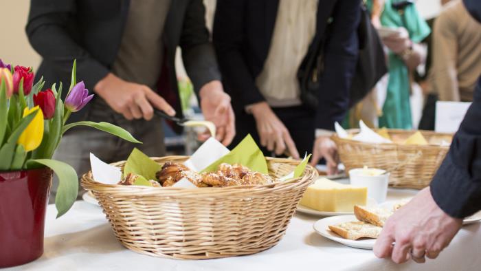Ett antal personer förser sig från ett bord med bröd, ost och bullar. En färggrann tulpanbukett pryder bordet till vänster