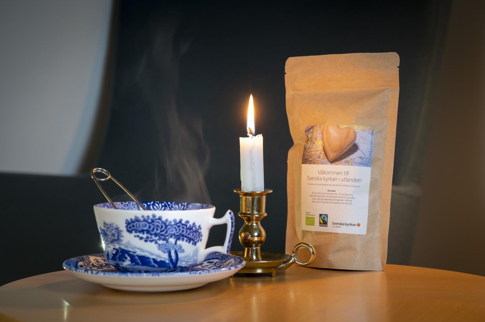 En rykande tekopp, ett tänt stearinljus och en påse med te från Svenska kyrkan står på ett bord.