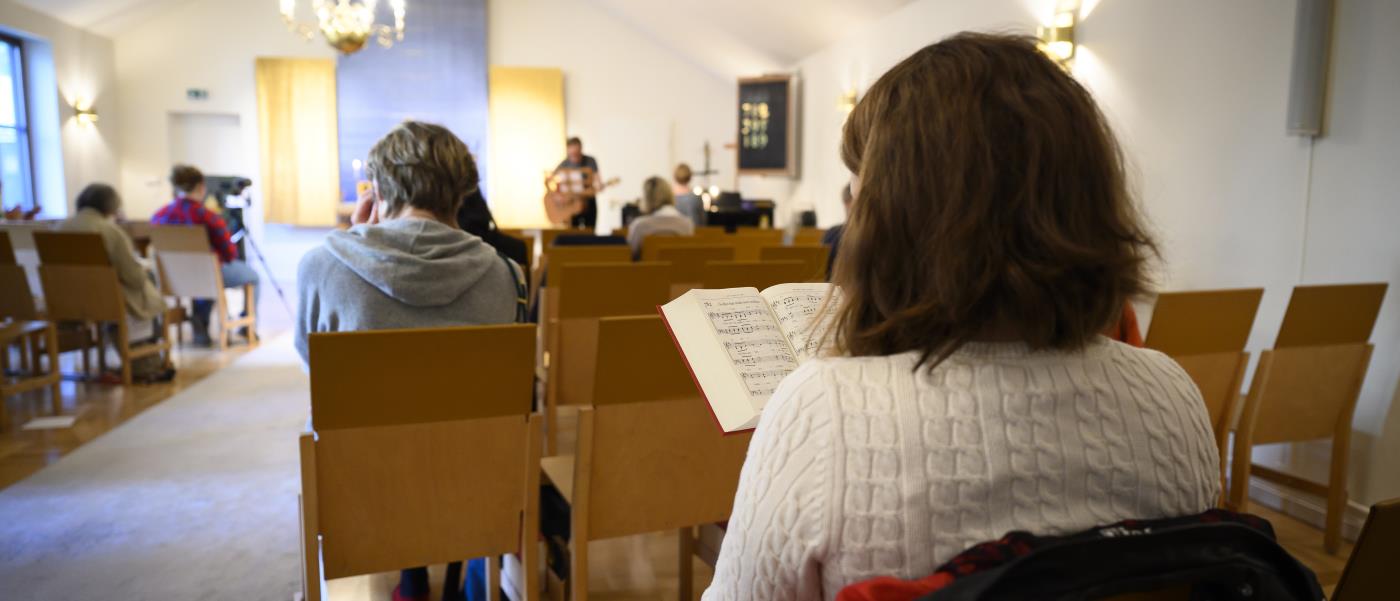 En kvinna sitter i en kyrkbänk med en psalmbok i handen. Flera besökare sitter på raderna framför.