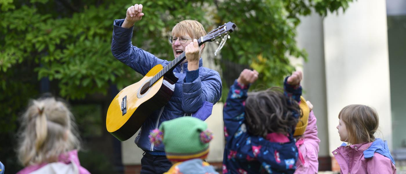 En kvinna står utomhus och spelar gitarr för en grupp barn.