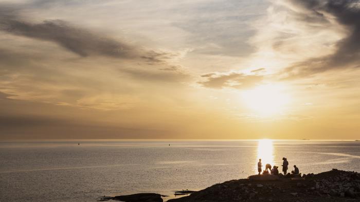 På en klippa vid havet har en grupp personer slagit sig ner för att se på solnedgången.