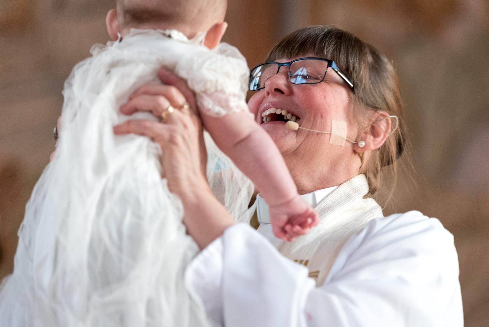 Kvinnlig präst håller leende upp ett dopbarn i luften.