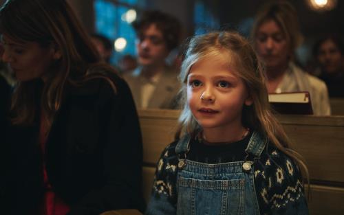 En blond liten flicka i snickarbyxor och stickad tröja sitter i en kyrkbänk. Andra besökare syns suddigt i bakgrunden.
