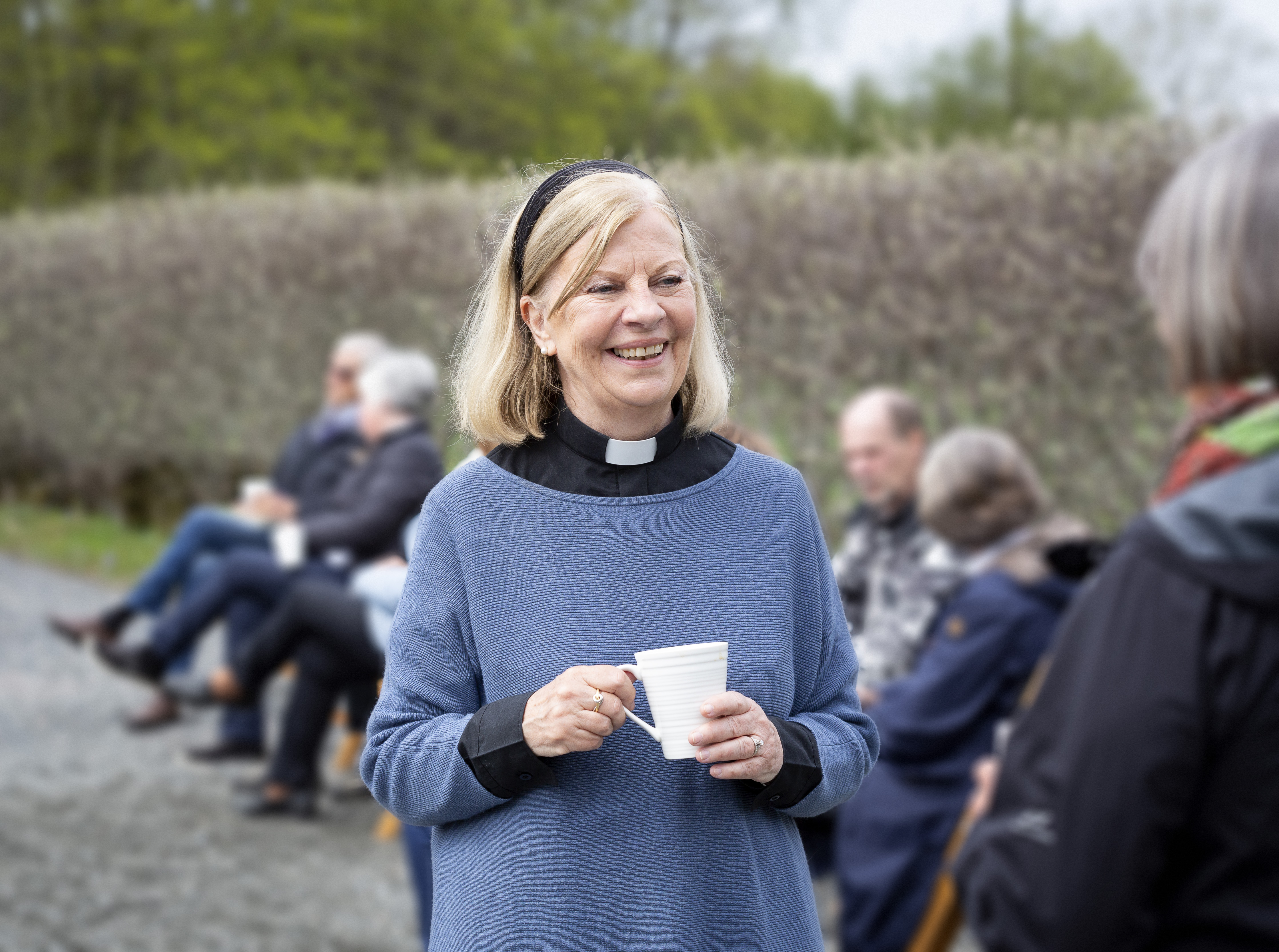 En kvinnlig präst står utomhus med en kaffemugg i handen och samtalar leende med någon.