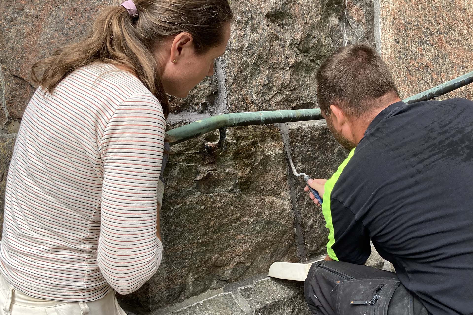 En kvinna och en man sitter på huk intill en mur av naturstenar. Mannen fyller i en fog med fogbruk mellan stenarna.