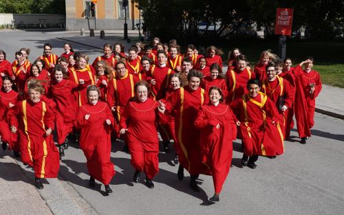 Uppsala domkyrkas ungdomskör springer glatt mot fotografen tvärs över gatan i sommarsolen, iförda röda körkåpor.