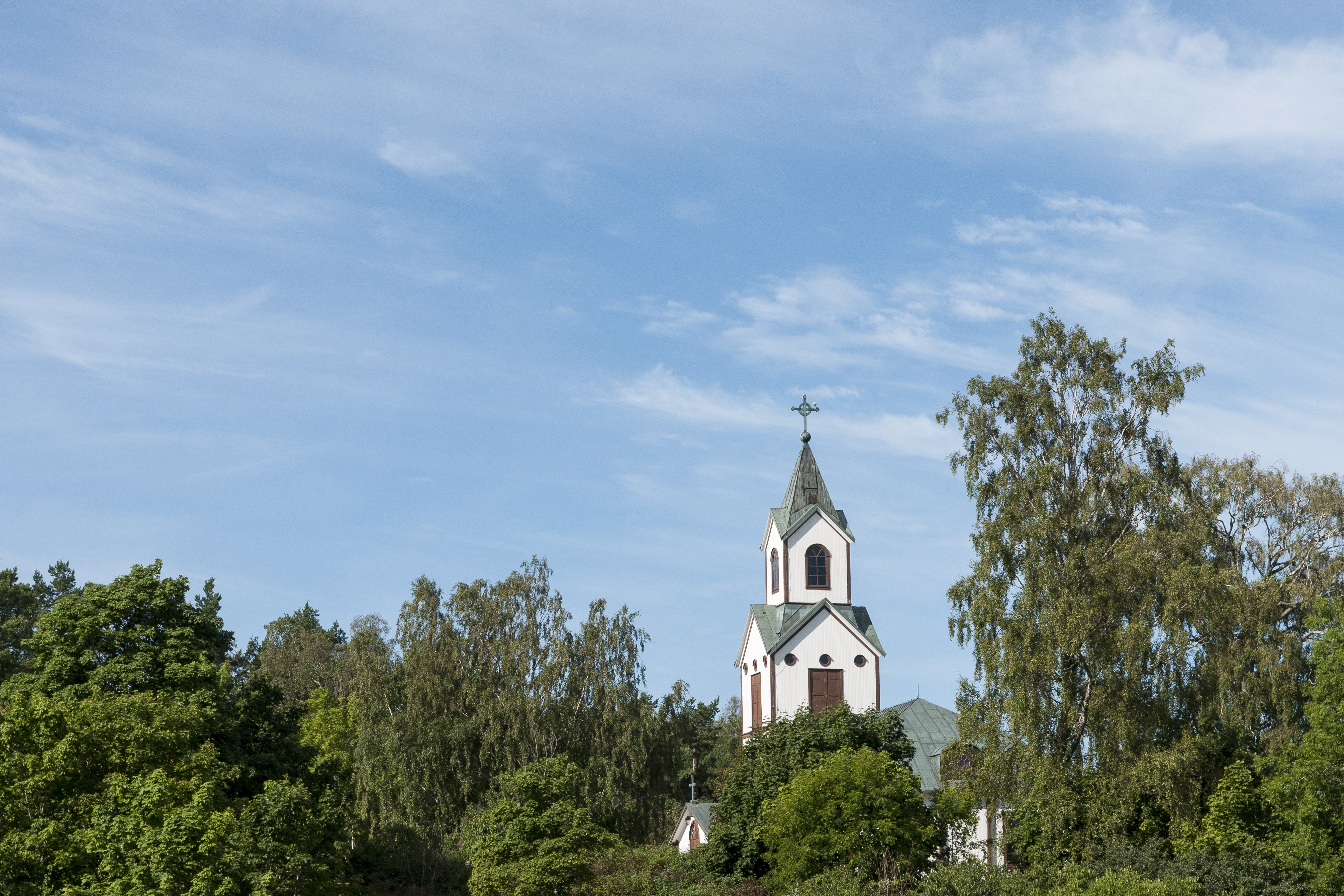 Ett kyrktorn sticker upp bakom trädkronorna mot blå himmel.