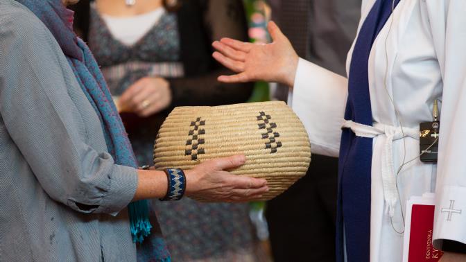 Kvinna i grå klänning överlämnar beige korg med kollekt till präst med utsträckt hand.