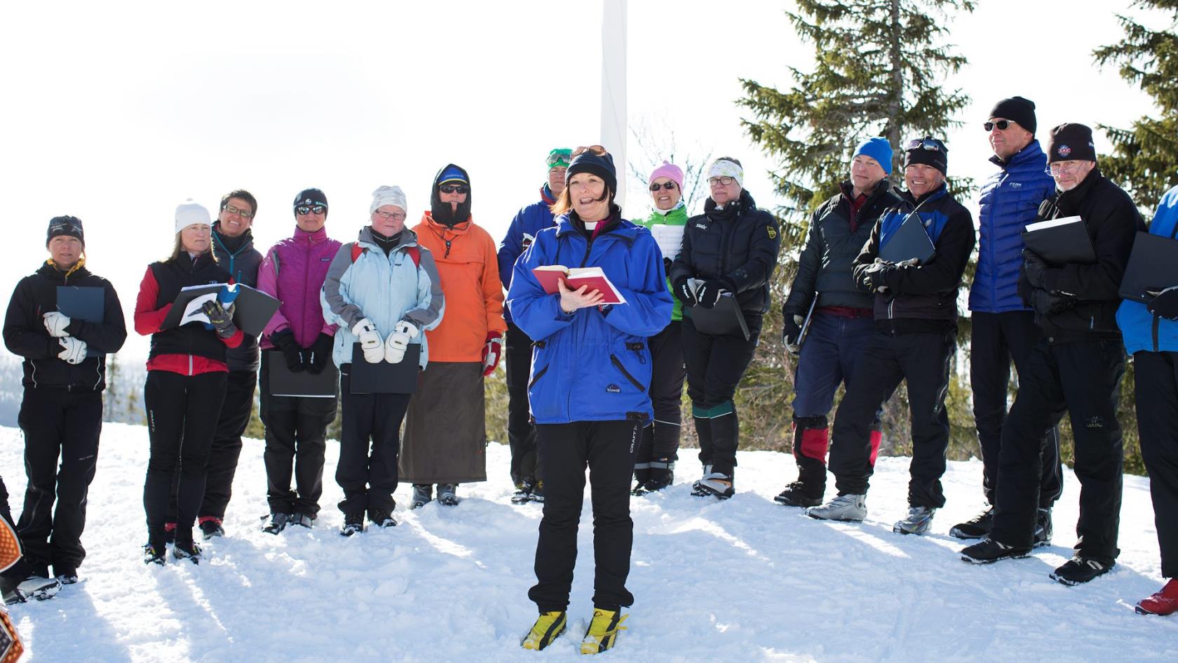 Utomhus en solig vinterdag. Prästen Karin Härjegård klädd i vinterkläder och skidpjäxor. Hon håller en psalmbok i sin hand och flankeras av ett tiotal personer också de klädda för vinter och skidåkning.