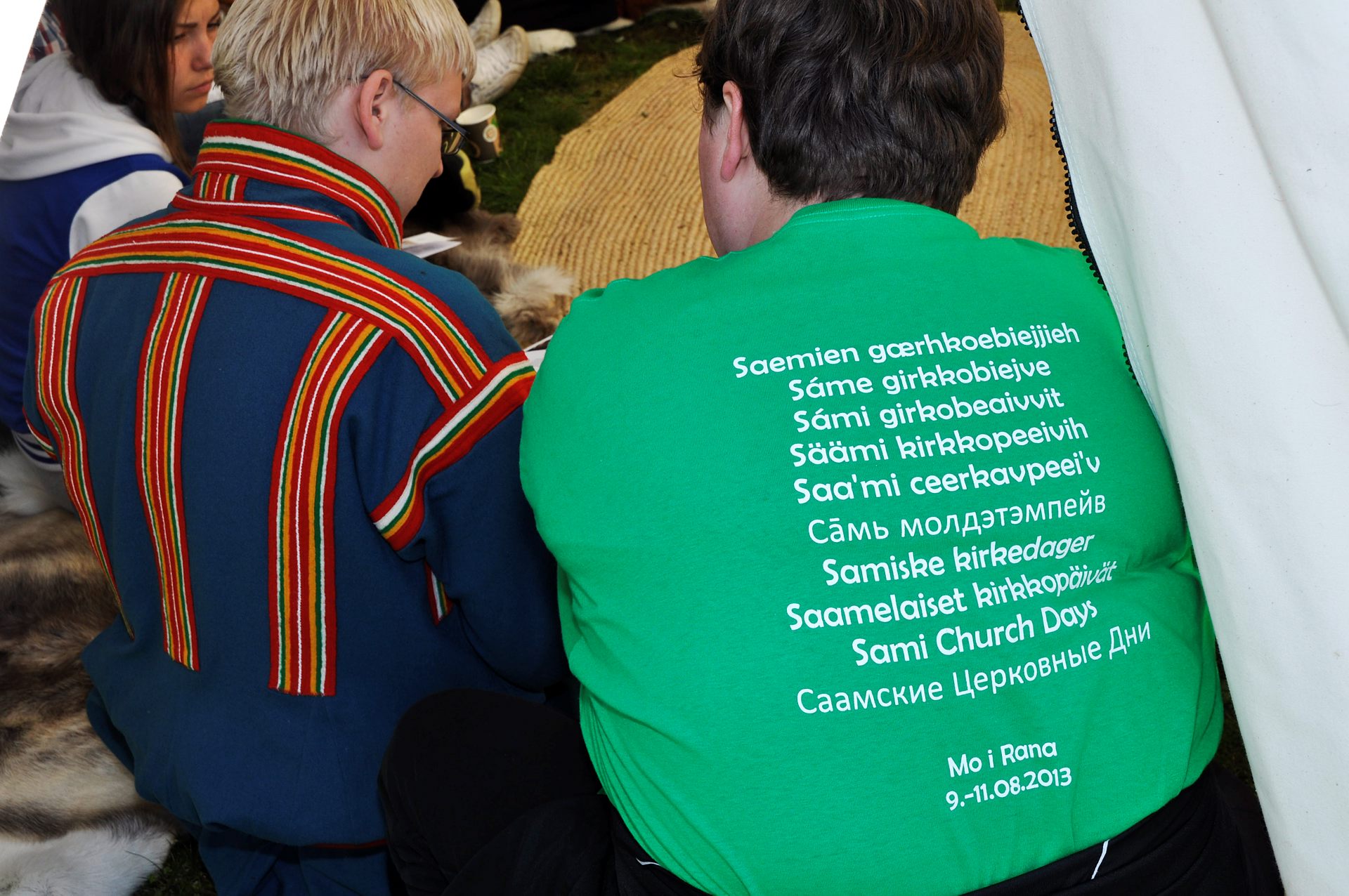 Två personer sitter bredvis varandra. Den ena har traditionell samisk dräkt. Den andra har en grön T-Shirt med texten "Samiska kyrkodagar" skrivet på et mängd olika språk.