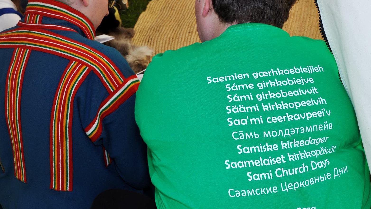 Två personer sitter bredvis varandra. Den ena har traditionell samisk dräkt. Den andra har en grön T-Shirt med texten "Samiska kyrkodagar" skrivet på et mängd olika språk.