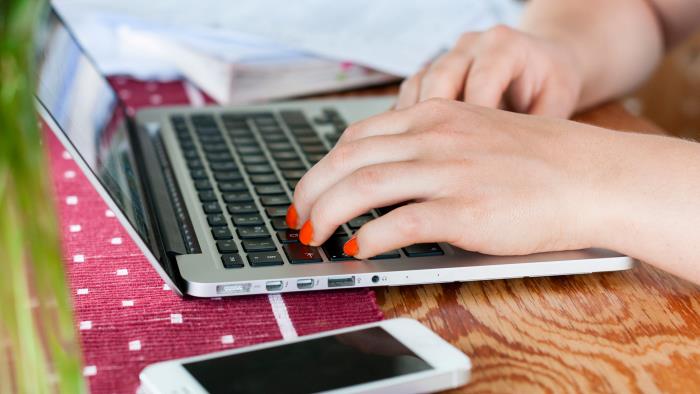 Två händer på en dators tangentbord, samt en mobil, en bok och några papper liggande på ett bord. 