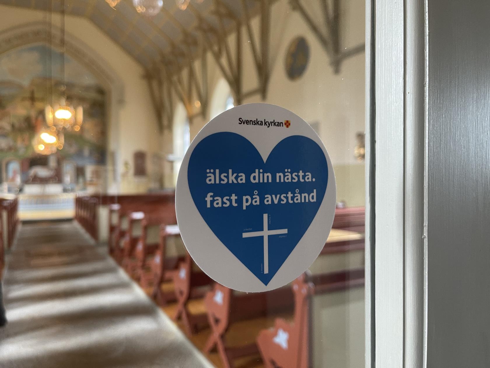 Ett klistermärkeshjärta med texten "Älska din nästa fast på avstånd" sitter på en fönsterruta i en kyrka