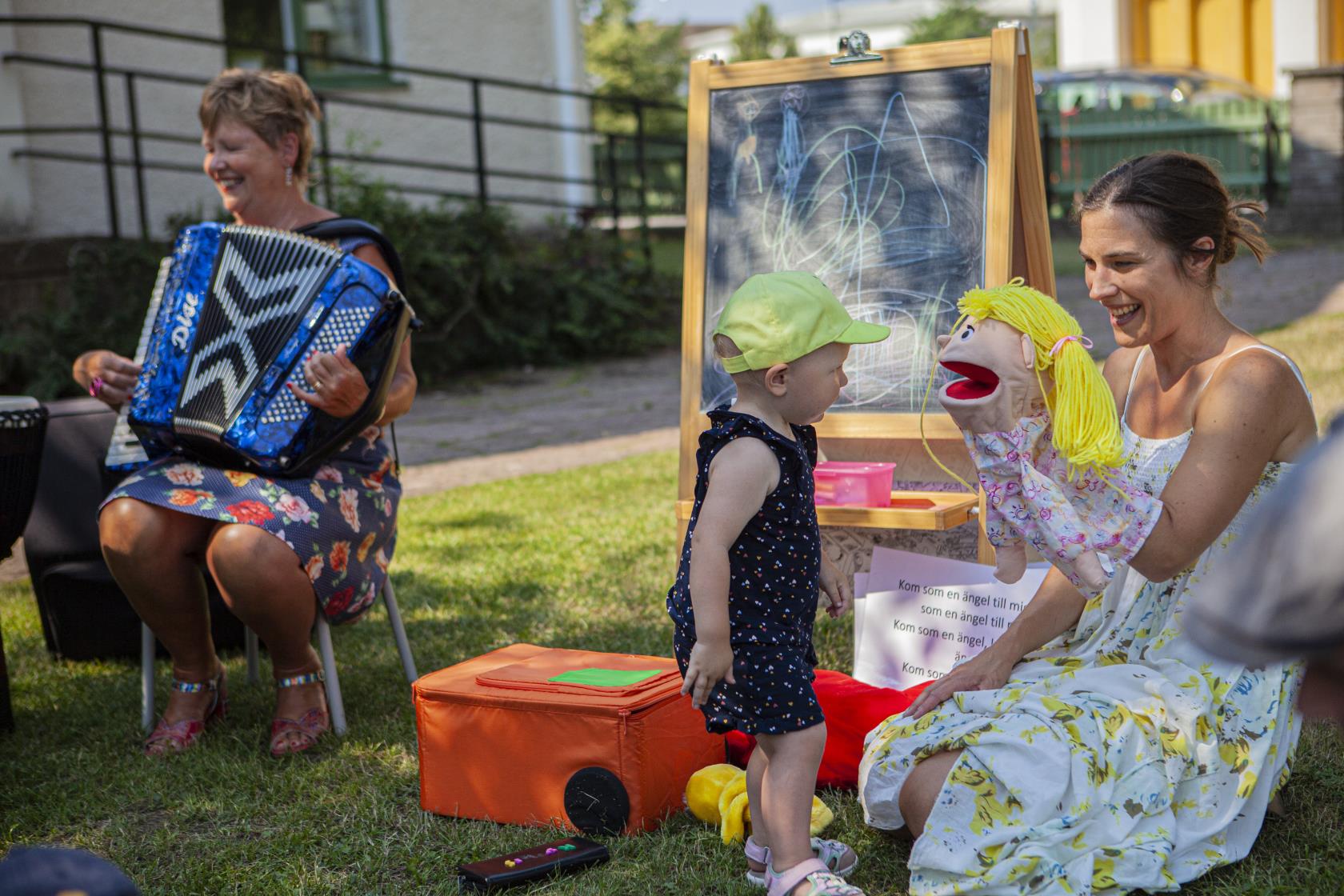 Handdockan Bibbi, barnledaren Julia och kantor Helen som spelar dragspel pratar med ett litet barn utomhus en sommardag