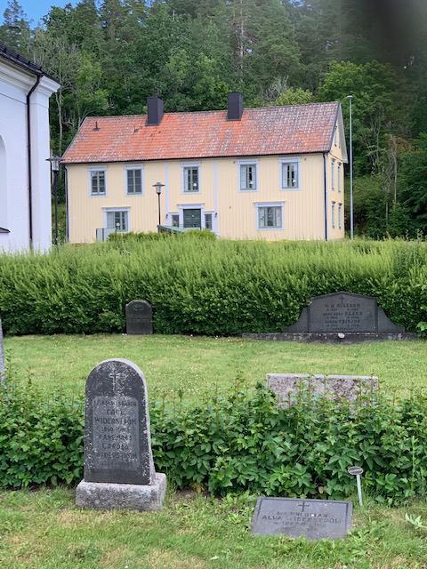 Gravsten över Alva Widerström och hennes fosterföräldrar på Västra Hargs kyrkogård. I bakgrunden ett gult hus, församlingshemmet.