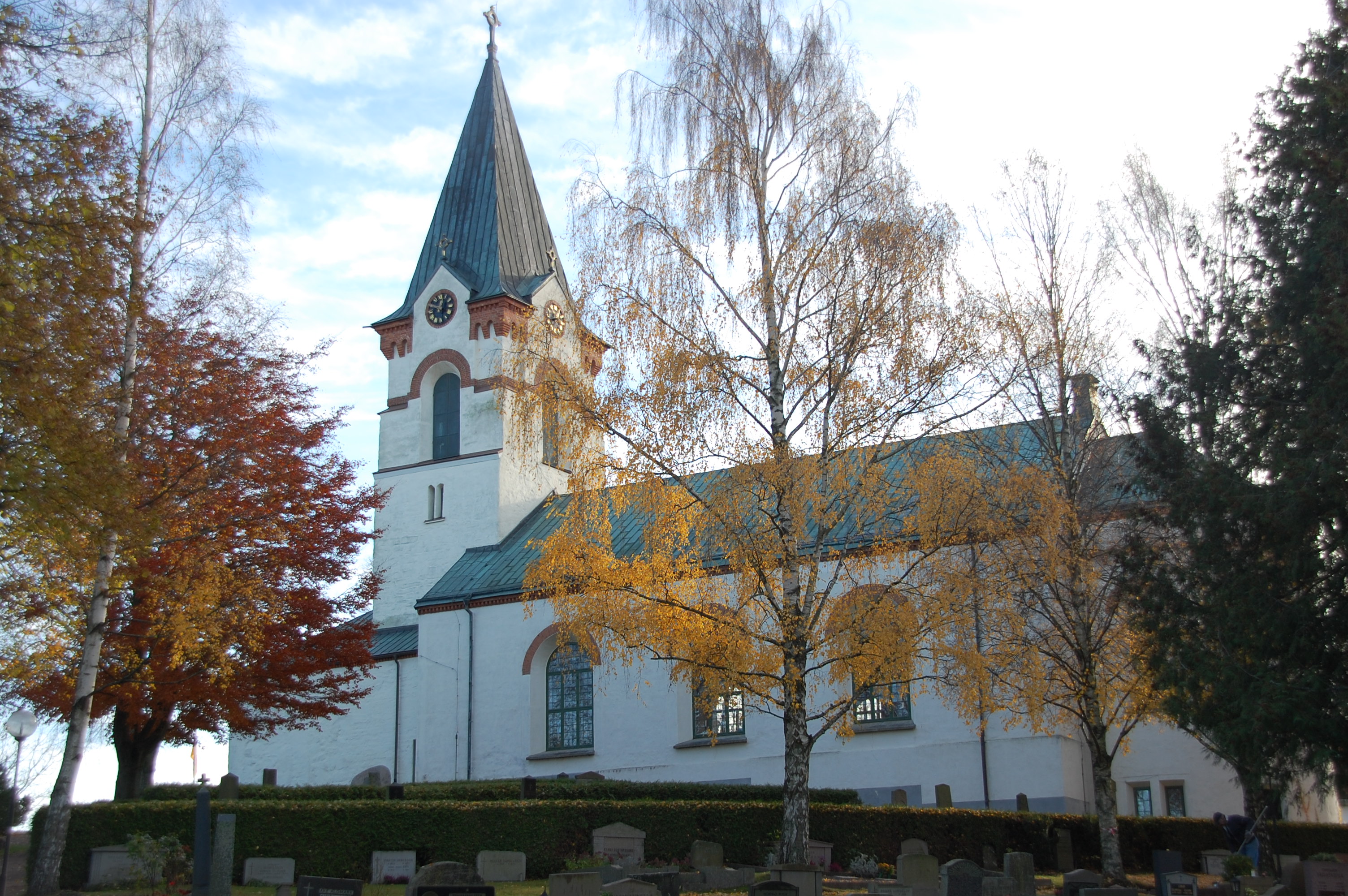 Ödeshögs kyrka, en vit kyrka med koppartak. Framför kyrkan finns gravvårdar på rad och träden skiftar i rött och gult.