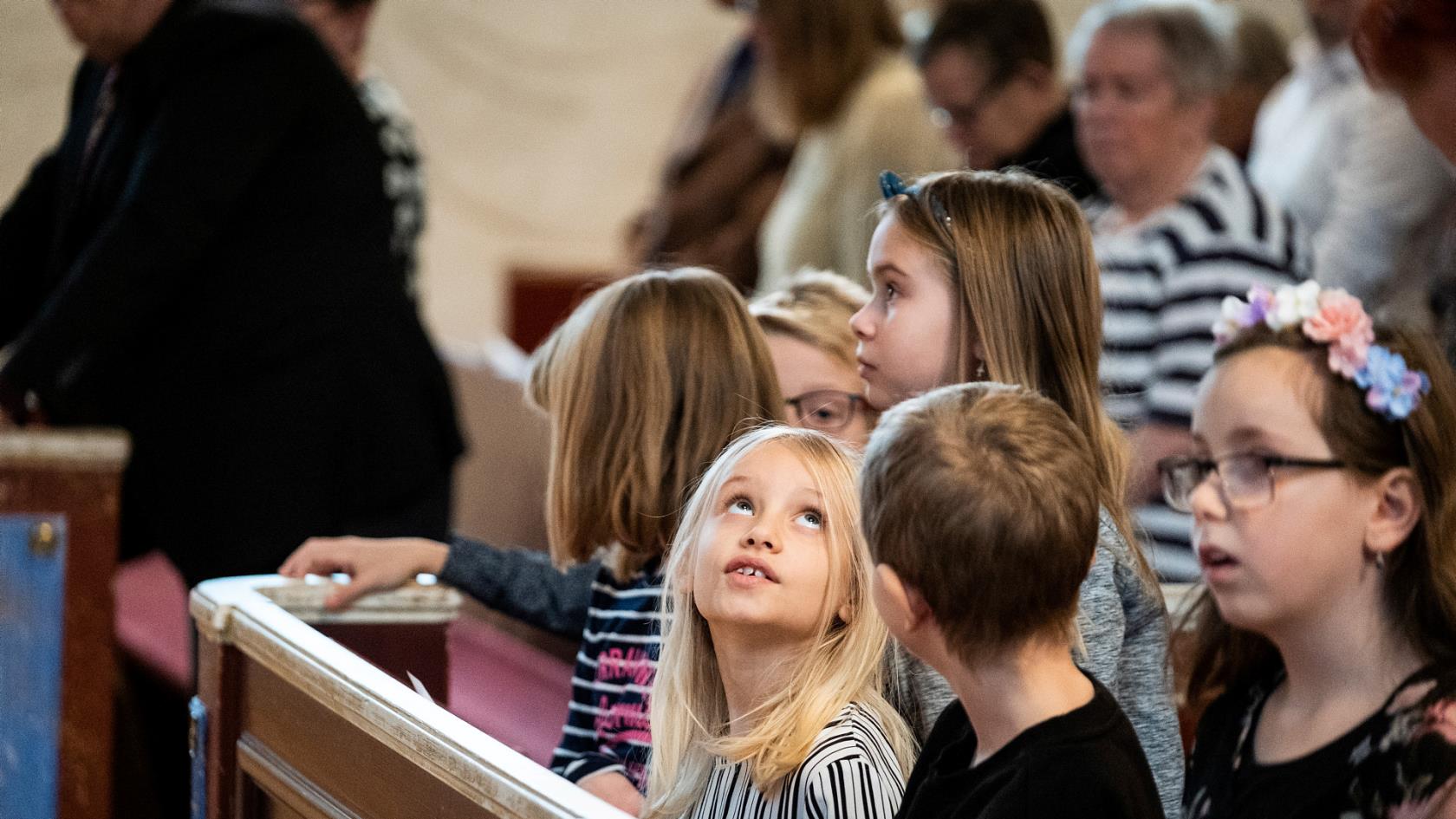 En flicka med blont hår tittar upp mot taket under en gudstjänst. Hon är omgiven av flera barn.