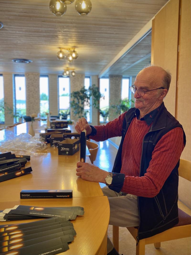 Volontären Janne Johansson sitter vid ett bord på Nathanaelgården i Tierp och paketerar kronljus i kartonger inför allhelgona. Janne har på sig en röd kabelstickad kofta men en svart väst över.